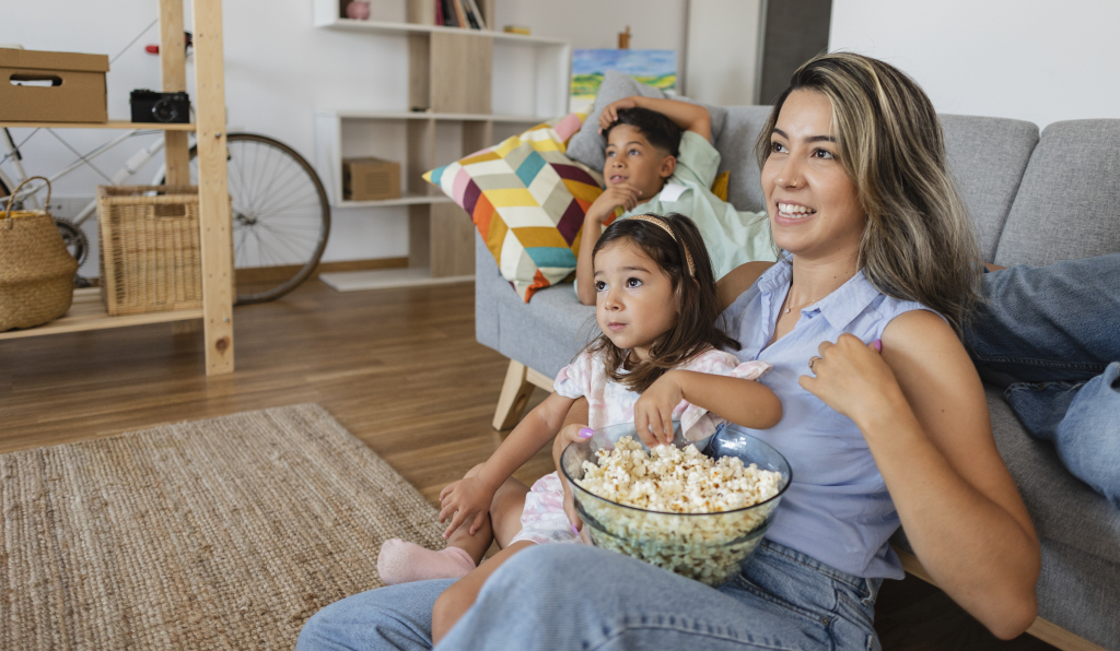 Beautiful young Hispanic mother and her children are watching TV, eating popcorn and smiling while sitting on the couch at home
